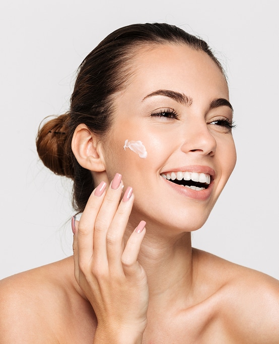 Femme souriante appliquant de la crème sur sa joue. Soin visage, peau saine, hydratation, beauté naturelle.