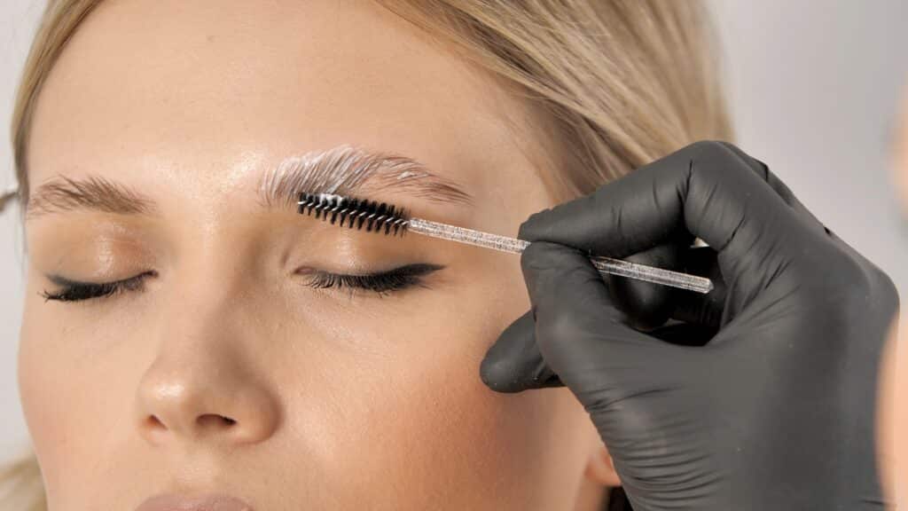 Femme aux yeux clos subissant un soin de lamination des sourcils. Une main gantée noire applique une crème blanche avec une brosse.