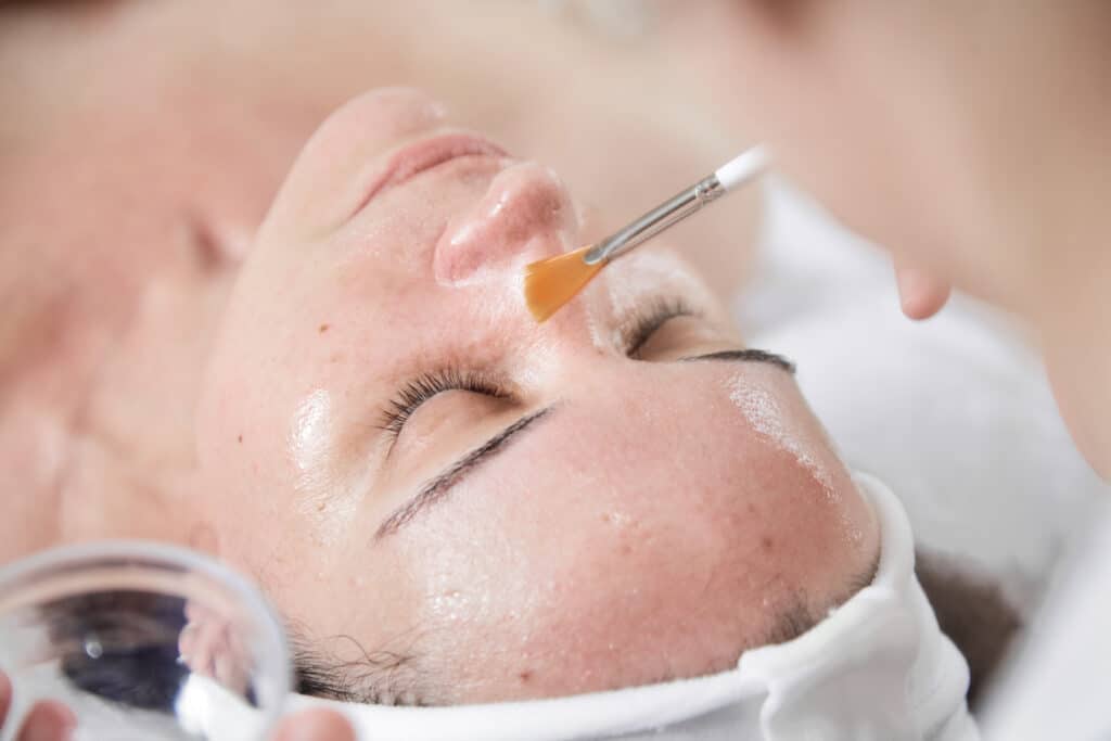 Esthéticienne appliquant un masque ou sérum à la brosse sur le visage d'une femme détendue, yeux fermés, lors d'un soin.